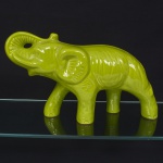 Escultura zoomórfica representando elefante, executada em cerâmica moldada, com corpo oco e acabamento vítreo de tonalidade verde uniforme. Modelagem tridimensional contínua, com volumes arredondados e tratamento estilizado da anatomia, evidenciando tromba alongada, orelhas amplas em relevo e dorso suavemente arqueado, cabeça apresenta olhos vazados, elemento funcional integrado à modelagem, base formada por quatro patas com bordas inferiores não vidradas, revelando a pasta cerâmica aparente. Peça sem marca aparente. Medidas: Alt.: 22 x Comp.: 38 x Larg.: 13 cm.