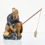 Escultura representando homem pescadondo na beira do riacho, executada em cerâmica moldada, com corpo oco e acabamento em vidrados policromos, apresenta barba bipartida e bigode alongado, cabelos presos em coque superior, traços fisionômicos acentuados e expressão serena, enverga túnica de mangas curtas em vidrado azul, cingida à cintura, com partes do corpo em vidrado marrom-claro; na mão esquerda segura chapéu típico, com decoração radial em relevo sob vidrado translúcido âmbar, mão direita segura vara de pesca, com conformação compatível para o encaixe da mesma; base integra a composição escultórica, modelada como afloramento rochoso, com vidrado de efeito marmorizado em tons escuros, evocando ambiente fluvial ou lacustre; apresenta orifício circular na base, típico de processos de queima e ventilação do corpo cerâmico. Base não vidrada, com marca em relevo CHINA e inscrição numérica/incisa adicional, indicando produção seriada, compatível com manufatura decorativa chinesa do século XX, voltada ao mercado ocidental, inspirada na iconografia tradicional do pescador como símbolo de simplicidade, trabalho e sabedoria. Medidas: Alt.: 13,5 x Comp.: 8,5 x Larg. 8 cm.