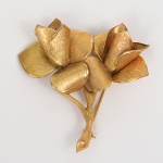 Broche floral em metal dourado, representando ramo com flores estilizadas, composto por múltiplas pétalas conformadas individualmente, com acabamento escovado acetinado e bordas polidas. Estrutura formada por haste central sinuosa, da qual partem os elementos florais articulados, flores apresentam pétalas côncavas sobrepostas, com nervuras finamente marcadas por escovamento direcional, conferindo contraste entre superfícies foscas e áreas de brilho. Verso com alfinete horizontal e fecho de segurança, integrado à estrutura metálica.Medidas: Alt.: 5,5 x Comp.: 5,5 cm.