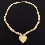 Colar composto por contas esféricas, ovais e cilíndricas em osso natural, de coloração marfim, perfuradas e montadas em fio contínuo, organizadas de forma simétrica ao longo do eixo central. A composição intercala elementos metálicos espaçadores, incluindo contas circulares estampadas com motivo floral em relevo e aneletes metálicos estriados, distribuídos em pares. Na extremidade inferior, apresenta pingente em formato de coração, confeccionado em osso maciço, com perfuração superior simples e suspensão por argola metálica. O fecho é do tipo rosqueável cilíndrico metálico, em duas partes, com extremidades estriadas. Produção compatível com a segunda metade do século XX, associada à joalheria artesanal de inspiração étnica. Estado de conservação bom, com sinais de uso compatíveis com a idade, pátina nos elementos metálicos e marcas naturais do material orgânico, sem perdas aparentes.Medida fechada: Comp.: 25 cm.(contando o pingente)