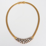 SWAROVSKI. Colar rígido articulado em metal dourado, feitio flexível tipo cobra (snake chain) de seção circular, terminado em fecho de encaixe articulado com trava central, assinado Swarovski. Na parte frontal, apresenta elemento central semicircular aplicado, em metal com acabamento contrastante, decorado por motivos geométricos vazados em padrão zigue-zague, intercalados por aplicações de cristais lapidados incolores cravados em superfície contínua, formando faixa decorativa de destaque. O reverso do elemento frontal é liso, com acabamento metálico uniforme. Apresenta discretos sinais de uso e desgaste superficial compatíveis com a idade, mantendo integridade estrutural e cravações íntegras. Medida fechada: 15,5 x 11,5 cm.
