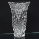 Vaso em vidro incolor, obtido por moldagem prensada, apresentando corpo obcônico, base circular escalonada e boca circular ondulada, superfície externa estruturada por painéis verticais facetados, intercalados por motivos em relevo compostos por cachos ovais em arranjo piramidal e rosáceas radiais de simetria axial, região inferior marcada por gomos alongados convergentes, culminando em estrela geométrica centrada, borda superior definida por perfil sinuoso contínuo, interior decorado por relevos geométricos repetitivos. Medidas: Alt.: 20 x Diam.: 12,5 cm.