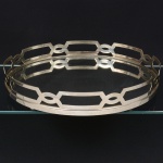 Elegante bandeja art déco, de formato oval, constituída por centro em vidro espelhado, alojado em estrutura metálica vazada, aro perimetral formado por faixa contínua com elementos geométricos interligados, apresentando recortes ovais e poligonais dispostos em sequência regular, apoiado sobre quatro pés metálicos hemisféricos, fixados à face inferior do vidro, garantindo afastamento da superfície de apoio, borda do espelho encaixada por pressão na moldura. Medidas: Alt.: 7 x Comp.: 38 x Larg.: 30 cm.