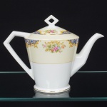 NORITAKE, Nagoya - Japão(Fundada em 1904 pelos irmãos Ichizaemon e Toyo Morimura). Bule para chá da década de 1950 em porcelana japonesa, estilo Art Déco; esmaltagem vítrwa na cor branca, bojo obcônico assentado sobre base circular, asa lateral estilizada, parte superior e tampa circunda por faixa de tonalidade marfim, dotada de decoração floral policromada por decalque, organizada em painéis centrais ovais e faixa perimetral contínua, acompanhada por elementos fitomórficos estilizados em azul e dourado. Filetes metálicos dourados delimitando tampa e campo decorativo superior, puxador de formato losangular, bico vertedor curvado. Marca da manufatura no verso da base. Medidas: Alt.: 18 x Comp.: 24 x Larg.: 13 cm.