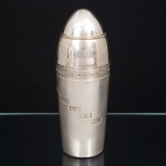 WOLFF. Raríssima coqueteleira em metal espessurado à prata, confeccionada com formato de projétil, apresenta duas tampas, uma para colocação das bebidas, outra para dispensar; possui dois copos, sendo o de dentro giratório que muda a escala de ingredientes conforme o nome do drink, que se apresentam em placas no copo de fora, constando neste exemplar: Alexander, Bacardi, Between The Sheets, Bronx, Clover Club, Dry Martini, Dubonnet, Gin Ryckey, Manhattan, Old Fashioned, Orange Blossom, Palm Beach, Sid Car, Tom Collins e Whisk Sour. Marca da manufatura e numeração puncionada ao fundo. Apresenta desgastes e sinais de uso e oxidação. Med.: Alt. 25,5 x Diam. 10 cm.