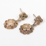 JOIA. Prata. Par de brincos confeccionados em prata, compostos por pino reto com tarraxa de pressão e elemento suspenso por argola articulada. Parte inferior formada por motivo floral estilizado, de planta circular, estruturado por núcleo central em relevo e elementos periféricos dispostos radialmente, ornamentação composta por aplicações de pequenas inserções lapidadas de pequeno formato(apresenta algumas perdas), em engaste fechado, distribuídas ao redor do motivo central. Superfície com trabalho de cinzelagem. Falta uma tarracha. Não possui contraste aparente. Porém, foi submetido ao teste. Medida: 2 cm. Peso: 3 gramas.