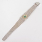 Relógio de pulso feminino, da marca HINA, movimento 17 jewels, caixa circular em metal branco, vidro convexo, mostrador circular de coloração verde, marcadores lineares aplicados e ponteiros centrais. Conjunto frontal ornamentado por pedras incolores lapidação brilhante, dispostas radialmente ao redor do mostrador, fixadas em engastes individuais. Pulseira integrada em malha metálica milanesa, de seção retangular, com fecho dobrável articulado. Coroa lateral estriada para ajuste. Verso da caixa em aço, marcada STAINLESS STEEL BACK, indicando fundo em aço inoxidável. Não está funcionando. Medida aberta: Comp.: 20 cm.