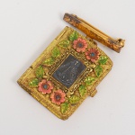 Relicário em formato de livro confeccionado em metal dourado, com dobradiça lateral e fecho de pressão. Verso com alfinete articulado para uso como broche e argola superior para suspensão em corrente. Anverso com moldura em relevo, decorada por elementos florais e folhagens aplicadas, com inserções policromadas. Campo central contendo imagem devocional de Nossa Senhora Aparecida, em representação frontal. Interior composto por duas faces espelhadas em relevo, com motivos simétricos de volutas, cruzes estilizadas e composição geométrica axial, obtidas por cunhagem em matriz. Execução por moldagem e estampagem mecânica. Peça destinada a uso devocional e ornamental. Apresenta pequenos sinais de oxidação, visíveis nas fotografias. Medida fechada: Alt.: 3,5 x Comp.: 3 cm.