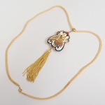 Colar composto por cordão têxtil trançado, de seção circular, com terminais metálicos e fecho tipo mola. Pingente central de formato recortado, apresentando representação figurativa de pavão em perfil, disposto sobre ramo vegetal, acompanhado por motivos florais. Decoração executada por esmaltação cloisonné, com campos de esmalte policromado separados por filetes metálicos aparentes. Contorno externo delimitado por filetes curvilíneos esmaltados. Parte inferior provida de borla pendente, formada por conjunto de fios metálicos fixados por terminal cilíndrico. Suspensão do pingente realizada por argola metálica articulada conectada ao cordão. Medidas: Comp.: 47 cm. Pingente: Alt.: 5,5 x Comp.: 4 cm.