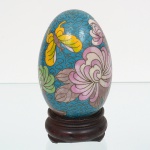 Ovo decorativo executado em metal esmaltado, decorado pela técnica cloisonné, com campos de esmalte policromado delimitados por filetes metálicos. Decoração composta por motivos florais estilizados, com flores de pétalas sobrepostas, folhagens lanceoladas e elementos fitomórficos curvilíneos, dispostos sobre fundo esmaltado contínuo com padrão repetitivo de traços sinuosos. Parte inferior provida de orifício circular central, compatível com suporte. Acompanha base circular em madeira, de perfil escalonado, de interio côncavo. Medidas: Alt.: 9 x Diam.: 6 cm.
