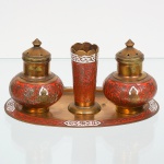 Conjunto de mesa composto por um(01) saleiro, um(01) pimenteiro e um(01) paliteiro, acompanhados de uma(01) bandeja, executado em metal amarelo com decoração em esmalte cloisonné, estilo orientalista. Bandeja de formato oval, com aba elevada, campo central liso e borda decorada por faixa esmaltada vermelha com motivos fitomorfos contínuos, intercalada por cartelas geométricas esmaltadas em branco com padrão estilizado. Saleiros de corpo globular, assentados sobre base circular, providos de tampa cônica removível com perfurações funcionais, decorados por campo esmaltado vermelho com arabescos lineares e motivo figurativo central em reserva, representando busto estilizado em nicho arqueado. Paliteiro central de corpo obcônico, com borda superior recortada em festões, decorado por campo esmaltado vermelho com traçado sinuoso contínuo e faixa superior em esmalte branco com padrão vazado. Medidas: Saleiros: Alt.: 6 x Diam.: 4,5 cm; Paliteiro: Alt.: 5,5 x Diam.: 2,5 cm; Bandeja: Comp.: 12,5 x Larg.: 8,5 cm.