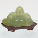 Escultura representando Budai, personagem da tradição chinesa, executada em material pétreo translúcido de tonalidade verde clara, compatível com jade ou serpentina, entalhada em bloco único. Figura representada sentada, com cabeça calva, fisionomia sorridente, olhos semicerrados, abdômen proeminente, vestes estilizadas com pregas incisas e braços apoiados lateralmente, integrados ao corpo escultórico, apresenta trabalho de entalhe manual, com modelagem contínua, volumes arredondados e acabamento por polimento, evidenciando translucidez irregular e variações naturais do material. Assentada sobre base independente em madeira, de coloração vermelha. Medidas: Alt.: 5 x Comp.: 9,5 x Larg.: 4,5 cm.