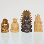 Conjunto composto por quatro(04) esculturas de iconografia sino-budista, executadas em materiais distintos, representando figuras tradicionais do panteão religioso e filosófico oriental: a) Buda sentado com halo  Figura em posição meditativa, assentada sobre base em forma de lótus. Corpo em liga metálica de tonalidade escura, com halo posterior dourado, recortado em raios estilizados e inscrições em caracteres chineses em relevo no reverso. Técnica de fundição em molde, com posterior acabamento manual e aplicação de pátina. Medidas: Alt.: 7 x Comp.: 4 x Larg.: 3,5 cm; b) Imortal/erudito taoísta  Escultura de personagem barbado, portando bastão, vestes longas e chapéu cerimonial, executada em resina de coloração marfim, entalhada em bloco único. Técnica de talha mecânica ou moldagem, com acabamento polido fosco. Medidas: Alt.: 6 x Comp.: 3 x Larg.: 3 cm. c) Budai (Buda Sorridente)  Representado sentado, com abdômen proeminente, sorriso acentuado e mãos segurando esfera simbólica, sobre base circular estriada. Executado em resina de tonalidade marfim, com modelagem em molde e posterior acabamento superficial. Medidas: Alt.: 4,5 x Comp.: 3 x Larg.: 3,5 cm. d) Buda dourado em trono de lótus  Pequena figura sentada em posição meditativa, com halo posterior integrado, executada em resina com pátina dourada. Medidas: Alt.: 4,5 x Comp.: 3 x Larg.: 3 cm.