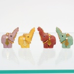 Conjunto composto por quatro(04) esculturas representando elefantes, executadas em resina moldada com pigmentação em diferentes tonalidades, apresentam modelagem figurativa simplificada, com corpo maciço, tronco elevado, orelhas laterais estilizadas, base plana e proporções compactas, adornadas com aplicações decorativas metálicas em tom dourado, compostas por elementos lineares fixados à tromba, argolas metálicas aplicadas à região do pescoço e plaquetas decorativas laterais. Medidas: Alt.: 5 x Comp.: 4 x Larg.: 2 cm; Alt.: 5 x Comp.: 5 x Larg.: 2,5 cm.