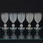 Elegante conjunto de taças de pé alto, para vinho tinto, em vidro prensado, diáfano e incolor; bojo oval moldado com sucessão de lonsagos relevados, padrão dito "bico de jaca", fuste haxagonal com anel intermediário, base circular de fundo estrelado. Composto de cinco(05) unidades. Medidas: Alt.: 14,5 x Diam.: 7 cm.