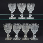 Elegante conjunto de taças de pé alto, para vinho do Porto, em vidro prensado, diáfano e incolor; bojo oval moldado com sucessão de lonsagos relevados, padrão dito "bico de jaca", fuste haxagonal com anel intermediário, base circular de fundo estrelado. Composto de sete(07) unidades. Medidas: Alt.: 13 x Diam.: 6,5 cm.