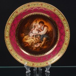 Prato decorativo em porcelana, corpo circular, cavado raso, com cena galante central representando casal em ambiente campestre, em composição inspirada na pintura europeia do século XVIII, com figuras trajando vestes de época, dispostas em atitude de cortejo, acompanhadas por cesto floral junto ao primeiro plano. Campo central em policromia, com modelado suave e gradações tonais, circundado por filete interno com motivos geométricos dourados aplicados a ouro, delimitando a transição para a aba, ornamentada por faixa magenta contínua, decorada com guirlandas entrelaçadas em dourado, ladeada externamente por banda creme com padrão floral e geométrico policromado, composta por flores estilizadas, estrelas e elementos lineares repetitivos. Borda externa com filete dourado contínuo. Marca da manufatura no verso. Medidas: Diam.: 25 cm.