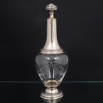 Exuberante licoreira francesa do início do século XX, executada em cristal soprado, de bojo oviforme assentado sobre base circular, decorada com guirlanda de ramos e folhas suspensas por laçarote, através de gravação a ácido; tampa, gargalo e base em prata francesa, relevada com faixa perimentral de motivo vegetalista. Apresenta contraste cabeça de Minerva, 1 Título, garantia do teor da prata do toque 0,950, usada a partir de 1838, e punção de ourives do mesmo período. Medidas: Alt.: 30,5 x Diam.: 12 cm.