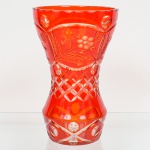 Vaso em cristal overlay vermelho, provavelmente de origem tcheca, apresentando camada externa rubi sobre cristal incolor. Forma obcônica com estrangulamento central e parte inferior alargada. Decoração executada por lapidação manual, composta por registros sobrepostos: faixa superior com motivos fitomórficos estilizados, com ramos, folhas e bagas; zona intermediária com padrão geométrico em retícula losangular; bojo inferior com reservas ovais e lapidação facetada. Borda superior recortada em ondulações regulares. Base espessa em cristal incolor, lapidada em estrela radial. Alt.: 23,5 x Diam.: 14,5 cm. Apresenta bicados ao redor da base.