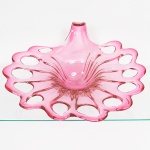 Belíssimo centro de mesa executado em vidro de Murano, tonalidade rosa, produzido por sopro em molde aberto. Corpo circular com centro côncavo, borda amplamente recortada em ondulações contínuas e vazados ovais regularmente espaçados ao longo do perímetro, aba conformada com canaletas verticais moldadas, dispostas de forma radial, convergindo para a região central, apresenta prolongamento curvo ascendente, integrado à borda, apresenta bolhas internas oriundas do processo de fabricação manual. Medidas: Alt. 17 x Comp.: 41 x Larg.: 37 cm.