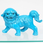 Escultura oriental executada em cerâmica esmaltada, tonalidade azul turquesa, representando Leão guardião chinês (Foo Dog) em posição de marcha, com cabeça voltada lateralmente, boca aberta e presas aparentes. Corpo robusto apoiado sobre quatro patas, cauda elevada e recurvada. Pelagem estilizada composta por mechas ondulantes em relevo contínuo, definindo juba, dorso e membros. Superfície integralmente revestida por esmalte vítreo de tonalidade azul-turquesa homogênea, olhos ressaltados com aplicação escura. Medidas: Alt.: 17 x Comp.: 26 x Larg.: 9,5 cm.