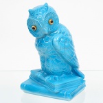 Escultura zoomórfica em cerâmica moldada, representando coruja em posição de repouso sobre base retangular escalonada composta por livros. Cabeça volumosa com olhos circulares salientes, compostos por inserções vítreas de coloração âmbar com pupilas escuras. Plumagem estilizada em relevo contínuo, com escamas e penas sobrepostas definindo cabeça, dorso, asas e peito. Superfície integralmente revestida por esmalte vítreo azul-turquesa homogêneo. Base vazada na face inferior, com abertura circular de queima, sem marca de fabricante visível. Medidas: Alt.: 18 x Comp.: 11 x Larg.: 10 cm.
