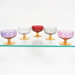 Conjunto composto de cinco(05) taças em vidro da década de 1960/70, bojo hemisférico em vidro translúcido nas cores rosa, incolor, vermelho e verde claro, apoiado sobre haste curta e base circular dotadas de profusa decoração em douradura. Medidas: