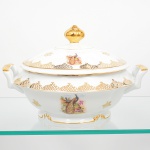 Porcelana Real, Mauá - SP(Fundada em 1943 por Fritz Erwin Schmidt). Belíssima sopeira em porcelana nacional da década de 1950/60. Esmaltagem vítrea na cor branca, decoração composta por cena com casal de pavões, pousados entre ramos, flores e folhagens, executada em estampagem policromada, realçada com generosos arremates em douradura, com motivo fitomórfico e treliçados em douração, distribuídos em reservas, e filete contínuo nas bordas; tampa abaulada de borda recortada, puxador em formato de fruto, alças laterais reviradas para cima. Marca da manufatura no verso. Med.: Alt. 18 x Comp. 28,5 x Larg. x Larg. 24,5 cm.