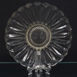 Travessa circular em vidro prensado, translúcido e incolor, com borda recortada em gomos regulares, decoração composta por estrias radiais facetadas partindo do centro em direção à borda, alternadas com planos triangulares formando composição geométrica repetitiva, centro com medalhão circular em relevo, decorado por estrela radial facetada, delimitado por anel frisado contínuo. Medida: Diam.: 32 cm.