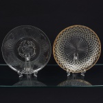 Dois pratos em vidro incolor, de formatos circulares distintos: Um apresenta aba larga com frisos radiais em relevo, cavidade central lisa, decorada por gravação floral composta por flor estilizada e ramos foliares. Borda com pequenos motivos vegetais espaçados. Fabricação por prensagem em molde, outro apresenta cavidade central com raios convergentes em relevo e aba trabalhada com padrão geométrico em losangos facetados, formando malha regular. Borda com filete dourado aplicado a frio. Fabricação por prensagem em molde. Medida: Diam.: 28 cm.
