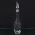 Garrafa em vidro incolor, composta por corpo soprado de perfil alongado, com bojo fusiforme e base circular plana. Gargalo estreito e alongado, com boca preparada para encaixe de tampa. Tampa facetada em vidro maciço, de forma prismática alongada, com rolha esmerilhada. Fabricação por sopro, com acabamento manual. Medidas: