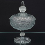 Compoteira em vidro prensado, ranslúcidoe  incolor, composta por bojo circular com tampa correspondente, fuste e base. Bojo e tampa com decoração em relevo geométrico em padrão losangular contínuo. Tampa encimada por pegador facetado integrado. Faixa horizontal em relevo delimitando a junção entre bojo e tampa. Fuste central com elemento nodular e base circular com relevo radial. Medidas: