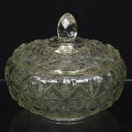 Bombonière em vidro prensado, translúcido e incolor, composta por recipiente e tampa, formato circular, perfil abaulado, assentada sobre anel de base plano, decorada por padrão geométrico repetitivo em estrelas facetadas, formando malha contínua em relevo, tampa decorada por malha geométrica de losangos e estrelas facetadas, com pegador central facetado de formato ovoidal, integrado ao corpo da tampa por base circular lisa. Medidas: Alt.: 15 x Diam.: 18,5 cm.