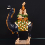 Grupo esculórico representando grupo de músicos montado sobre camelo com músicos (estilo Tang / inspiração sancai). Escultura executada em terracota vitrada policromada (sancai), representando um camelo bactriano com grupo de músicos montados no dorso. O animal exibe corpo modelado tridimensional, com patas robustas, pescoço arqueado e cabeça erguida; a superfície da peça é revestida com esmaltes coloridos em tons terrosos e variações de verde, âmbar e preto, aplicados em zonas, de acordo com a tradição de cerâmica vidrada chinesa (sancai  literalmente três cores), típica de artefatos funerários da dinastia Tang (618907 d.C.). O camelo serve de plataforma  uma pequena trupe de músicos ou viajantes  um motivo figurativo que aparece em esculturas cerâmicas chinesas associadas à vida em rotas de comércio, como a Rota da Seda, e à iconografia funerária que representava acompanhantes e servos (mingqi) do falecido no além-túmulo. A peça se apoia sobre base plana integrada, permitindo exposição em superfície horizontal. A modelagem demonstra variações de textura e volume concentradas no corpo do animal e nos detalhes das figuras, com interpretações estilizadas dos instrumentos e vestimentas. O acabamento sugere um objeto produzido de forma artesanal ou em pequena série inspirada em modelos arqueológicos antigos. Apresenta perda de uma perna de um dos personagens, e integrante que está em pé está com a cabeça colada. Medidas: Alt.: 52 x Comp.: 32 x Larg.: 15 cm.