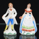 Duas(02) esculturas modeladas em porcelana esmaltada branca e decoradas manualmente com policromia. Represnetando casal, com figura masculina trajando casaca longa, colete e calções, com pintura em azul, amarelo e detalhes em dourado; postura ereta, braços junto ao corpo, apoiada sobre base integrada, e figura feminina trajando vestido longo de saia ampla, com aplicação de faixas decorativas em azul, verde e vermelho, corpete com pintura em vermelho e elementos florais estilizados. Medidas: Alt.: 10 x Comp.: 5,5 x Larg.: 3 cm; Alt.: 10,5 x Comp.: 5,5 x Larg.: 3,5 cm.