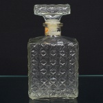 Garrafa executada em vidro incolor prensado,corpo de seção quadrangular, com ombros levemente chanfrados e gargalo curto com anel de reforço. Superfícies laterais integralmente decoradas por relevo geométrico repetitivo, composto por motivos estrelados facetados dispostos em padrão contínuo. Medidas: Alt.: 21 x Comp.: 11 x Larg.: 7 cm.