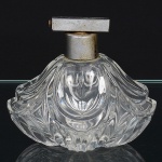 Frasco de perfume em vidro incolor prensado, corpo oblongo achatado, conformação lobada, com superfície moldada em relevo contínuo de volutas e nervuras curvas, convergindo para reserva circular central lisa na face inferior, base plana integrada ao molde, sem marca visível de fabricante, bocal cilíndrico curto, guarnecido por conjunto de pulverização metálico, com acabamento prateado, acionamento superior por alavanca basculante, sistema interno por tubo imerso(ausente). Medidas: Alt.: 10 x Comp.: 11 x Larg.: 7 cm.