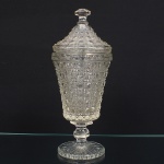 Grande compoteira executada em vidro prensado, translúcido e incolor, de bojo obcônico ricamente moldado com padrão geométrico, contendo losangos e octogonos relevo, fuste facetado sobre base circular de fundo estrelado, padrão decorativo replicado na tampa. Medidas: Alt.: 31,5 x Diam.: 13,5 cm.