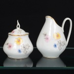 Porcelana Real, Mauá - SP(Fundada em 1943 por Fritz Erwin Schmidt). Uma(01) leiteira e um(01) açucareiro em porcelana nacional da década de 1960, esmaltagem vítrea na cor branca, decoração aplicada por estampagem policromada, com motivos florais estilizados compostos por flores radiais em tons de rosa e amarelo, acompanhadas por folhas em grisaille, distribuídos no corpo e na tampa, tampa abaulada com pega vazada elevada, moldada na própria porcelana, filetes dourados aplicados na borda superior do corpo e na aba da tampa, alça lateral curvada, bico vertedor ondulado. Marca da manufatura no verso. Medidas: Leiteira: Alt.: x Comp.: x Larg.: ; Açucareiro: Alt.: x Diam.: