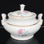 Porcellana Mauá, Mauá - SP(Fundada em 1937 por Fritz Erwin Schmidt / ativa até 1968). Sopeira em porcelana nacional da década de 1950/60; esmaltagem vítrea na cor branca, decorada com crisântemo e folhagens em estampagem policromada, predominando as tonalidades violeta, rosa, azul e verde; borda recortada em ondualções decorada com aplicações de filete dourado, circundada por volutas relevadas na própria pasta, e faixa contínua de folhagens aplicada em douradura, tampa ligeiramente abaulada encimada por puxador em forma de tulipa, alça laterais reviradas para cima. Marca da manufatura no verso. Medidas: Alt.: 19 x Comp.: 23,5 x Larg.: 22 cm.