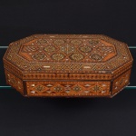 Caixa octogonal confeccionada em madeira, com tampa articulada por dobradiças metálicas e fecho frontal. Superfície externa integralmente decorada por marchetaria geométrica, composta por padrões simétricos de losangos, estrelas, faixas lineares e painéis poligonais, executados com lâminas de madeiras de diferentes tonalidades e incrustações claras, formando composição radial no tampo e frisos contínuos nas faces laterais, tampo apresenta campo central com motivo estrelado circundado por painéis geométricos repetidos, delimitados por molduras lineares,  laterais exibem painéis losangulares alternados, com incrustações triangulares e retangulares, mantendo coerência formal com o desenho do tampo. Interior em madeira lisa, sem forração, com estrutura montada por encaixes e colagens tradicionais. Necessita reparo na dobradiça, como mostrado nas fotografias. Medidas: Alt.: x Comp.: x Larg.: