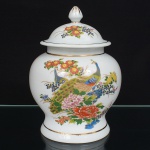 Pote de porcelana oriental do tipo ginger jar, com corpo bojudo, ombro pronunciado, boca circular e tampa campaniforme em forma de pináculo. Superfície esmaltada em branco, decorada em policromia sob e sobre o vidrado, com representação de aves estilizadas entre ramos florais e peônias, distribuídas no bojo e na tampa. Aplicação de frisos dourados na borda da boca, no ombro, na base e no remate da tampa. Base anelar. Fundo com inscrições em caracteres chineses pintados à mão, em disposição vertical. Técnica de pintura esmaltada (famille rose / famille verte combinadas, policromia oriental). Medidas: Alt.: 22 x Diam.: 16 cm. Apresenta um bicado na borda externa da tampa.