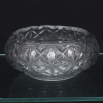 Centro de mesa em vidro prensado, translúcido e incolor, formato circular, de perfil baixo e abaulado, com borda suavemente recortada e base circular plana, decoração moldada composta por painéis geométricos estrelados em alto-relevo, formando rosetas de múltiplas pontas interligadas por facetas triangulares, distribuídas de maneira contínua ao redor do corpo. Interior decorado por estrela radial moldada no fundo, delimitada por anel circular em relevo. Aba superior com friso interno facetado, acompanhando o contorno da peça. Medida: Alt.: 9 x Diam.: 19 cm.