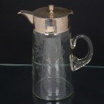 Jarra executada em cristal europeu, translúcido e incolor, decorada por lapidação manual com motivos fitomórficos distribuídos no bojo; corpo de perfil cilíndrico, alça curvada aplicada sobre a superfície, tampa em metal prateado, com sistema basculante, encimada por puxador central roscado, bico vertedor ondulado. Medidas: Alt.: x Diam.: