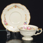 KPM - Krister Porzellan-Manufaktur, Waldenburg - Alemanha(Fundada em 1831 por Karl Kristen). Excepcional chávena para chá em porcelana de manufatura alemã, fabricada na polônia entre anos de 1930-1945, modelo Friederike, citado na Enciclopédia de Porcelana da Silésia Volume I. Série "Royal Ivory". Executado ao estilo renascentista. Esmaltagem vítrea de tom marfim, modelada com elementos fitomorfos em relevo, decorada com flores soltas em estampagem policromada, e generosos arremates de frisos em douraudura, chávena de bojo campaniforme, assente sobre base alta de formato circular e alça lateral em voluta, padrão decorativo replicado no pires e no prato. Marca da manufatura no verso. Meds.: Chávena: Alt. 6 x Comp. 12 x Larg. 10 cm. Pires: Diam. 15 cm.