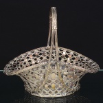 Art Krupp Metal, Bendorf - Alemanha(Fundada por Friedrich Krupp em 1811). Cesta confeccionada em metal prateado, fabricação alemã; formato circular, corpo vazado, confeccionado em trama entrelaçada de fios metálicos dispostos em padrão geométrico regular. Borda reforçada com aro liso aplicado. Alça superior em arco, formada por hastes múltiplas paralelas, reunidas no topo por amarra estriada. Base circular vazada, integrada à estrutura do corpo. Execução por técnicas de conformação, entrelaçamento manual e soldagem. Marca da manufaura puncionada. Medidas: Alt.: 21,5 x Comp.: 25 x Larg.: 18,5 cm. Apresenta desgastes no banho de prata.