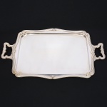 FRACALANZA - MODELO REGENCE. Grande bandeja confeccionada em metal espessurado à prata, c.1930-50, centro ligeiramente rebaixado, borda ondulada contornada por frisos e laços estilizados em relevo, obtidos através da técnica do cinzelado, padrão decorativo replicado nas alças laterais. Marca da manufatura puncionada ao fundo. Med.: Alt. 5 x Comp. 65 x Larg. 42 cm.