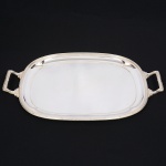 FRACALANZA. Belíssima bandeja confeccionada em metal espessurado à prata, formato retangular arredondado, centro rebaixado, borda relevada com fileira de frisos e fitas cruzadas, padrão "Croisé", ornato replicado nas alças laterais, com terminações cinzeladas em folhagens. Marca da manufatura puncionada no verso. Medidas: Alt.: 3 x Comp.: 52(de uma alça a outra) x Larg.:  33 cm.