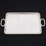 WOLFF. Elegante bandeja confeccionada em metal espessurado à prata, formato retangular de quinas arredondadas, centro ligeiramente rebaixado, borda percorrida por contorno relevado, alças laterais em "C". Marca da manufatura puncionada no verso. Medidas: Comp.: 46 x Larg.: 27,5 cm.