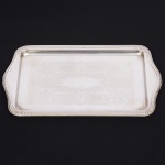 Elegante travessa para pães, ou bolo, confeccionada em metal espessurado à prata, formato retangular de abas laterais onduladas, borda contornada por sucessão de barras verticais em relevo, interrompidas por conchas nas quinas, centro ligeiramente rebaixado, lavrado com composição de motivo vegetalista. Med.: Comp. 36 x Larg. 20 cm.